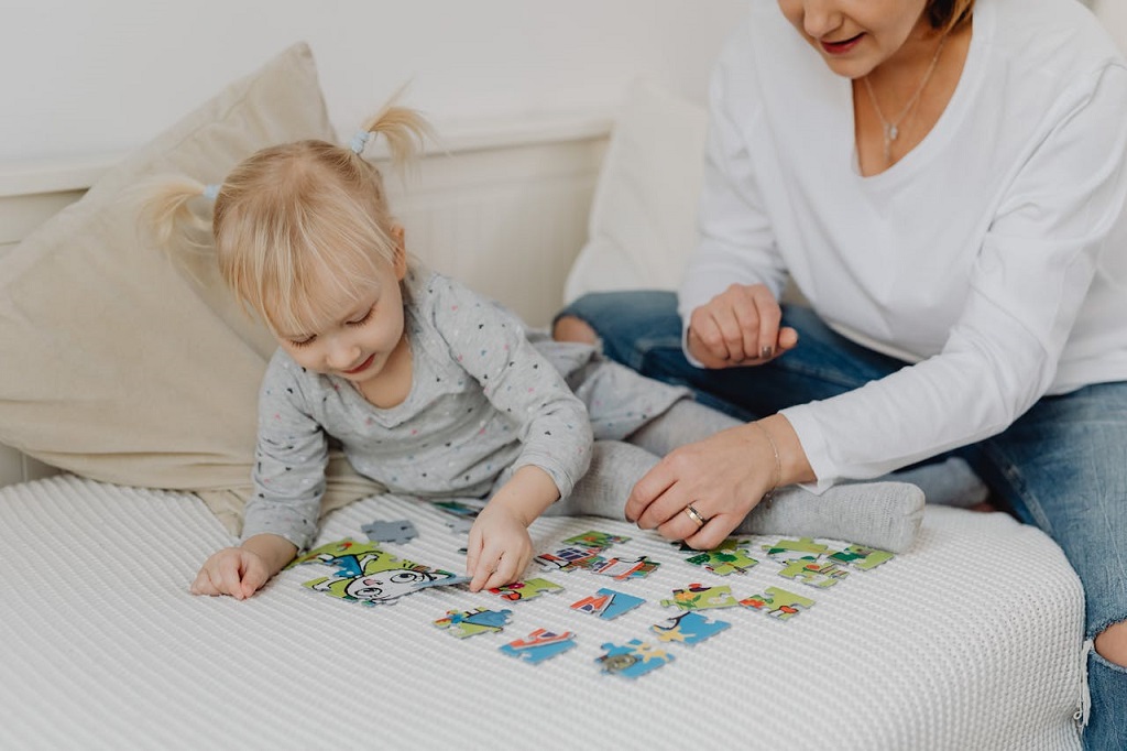 Devojčica i mama slažu šareni dečji slagalica-puzzle na krevetu, kao jednu od najboljih društvenih igara za decu